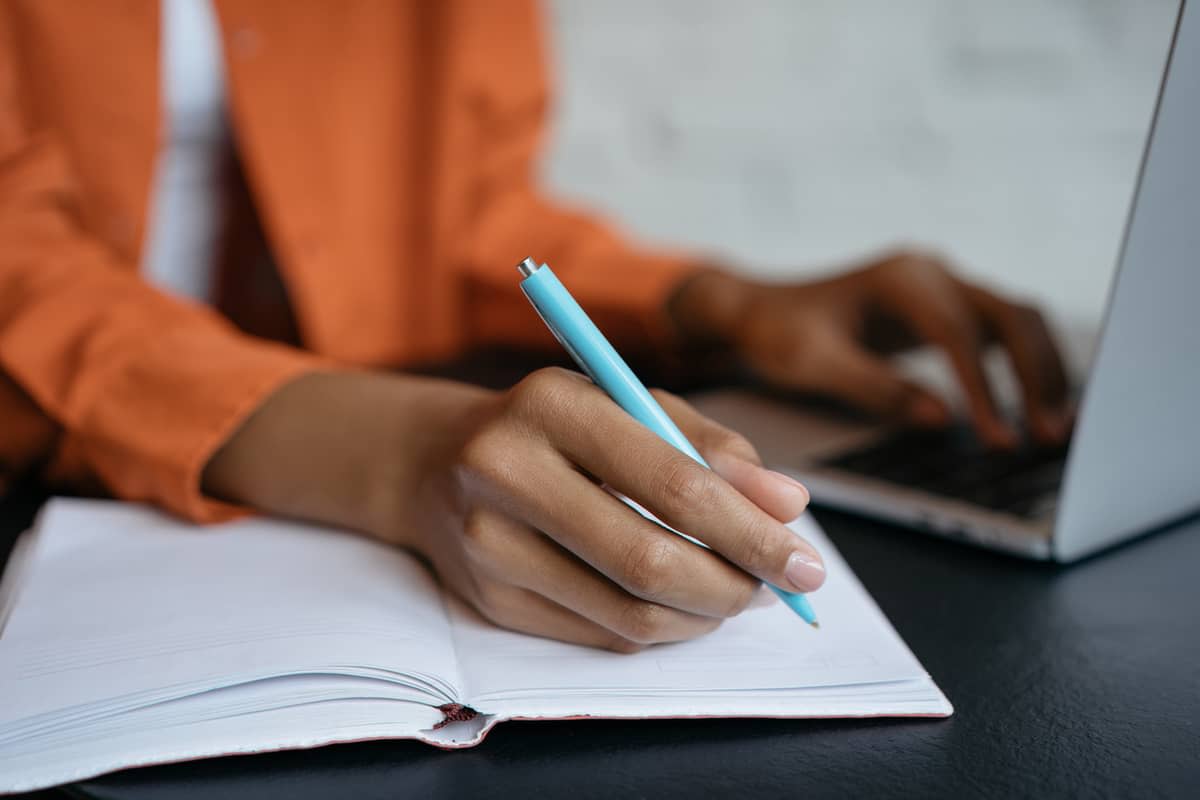 A woman holding a pen over a notebook while also working at a laptop