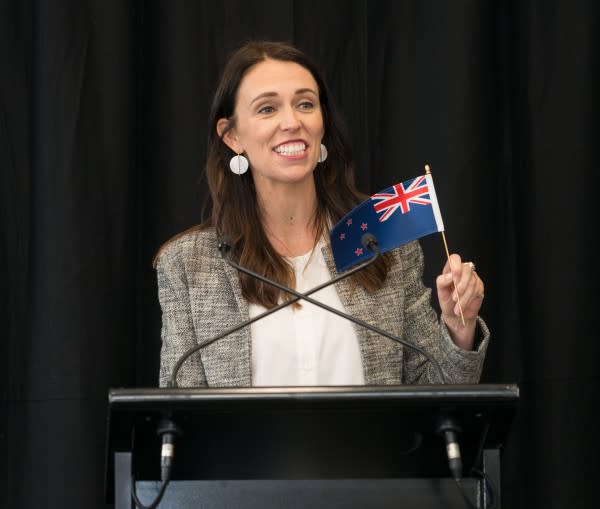 New Zealand Prime Minister Jacinda Ardern at a lectern, holding a small NZ flag. 