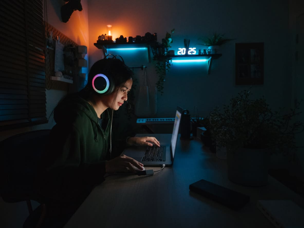 A girl sitting in a dark room illuminated by blue light, wearing headphones and using a laptop