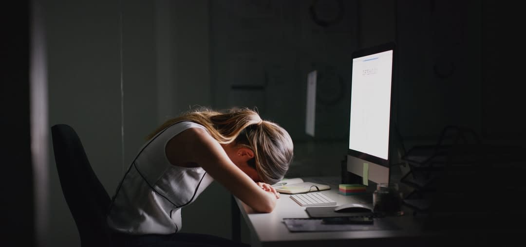 A woman sits at her desk in front of her computer with her head in her hands.