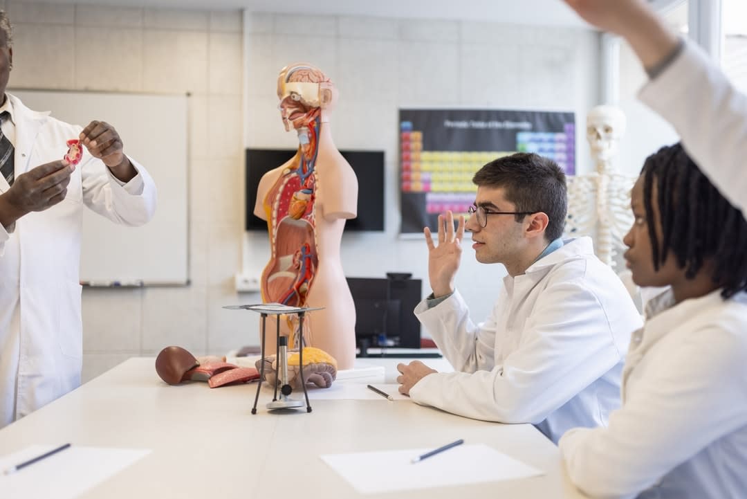 Students in lab coats raise hands to question a lecturer with an anatomical dummy.