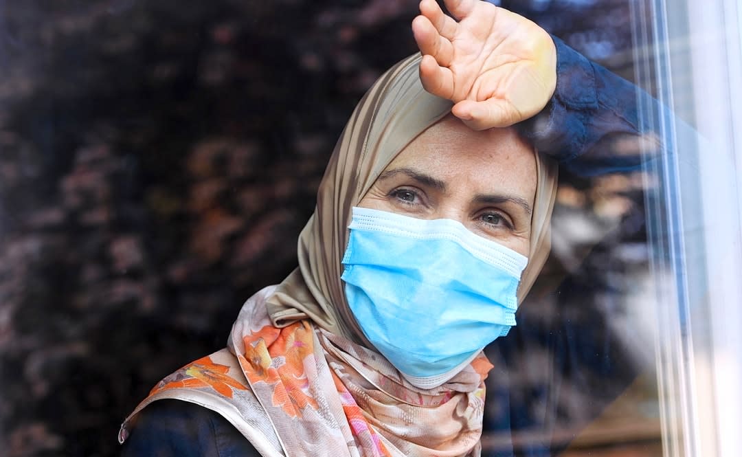 A woman with a headscarf and mask leaning against and looking out a window