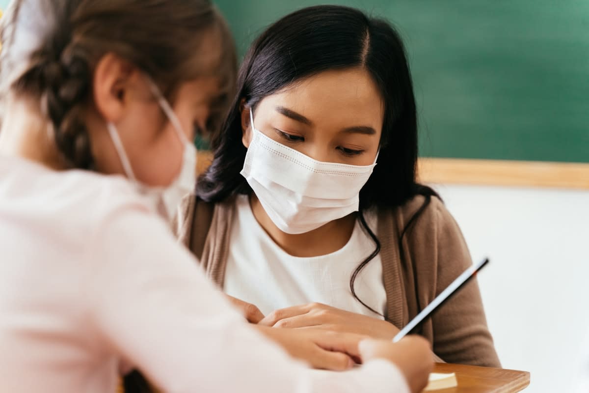 A masked woman teacher helping a young girl with her schoolwork in a classroom