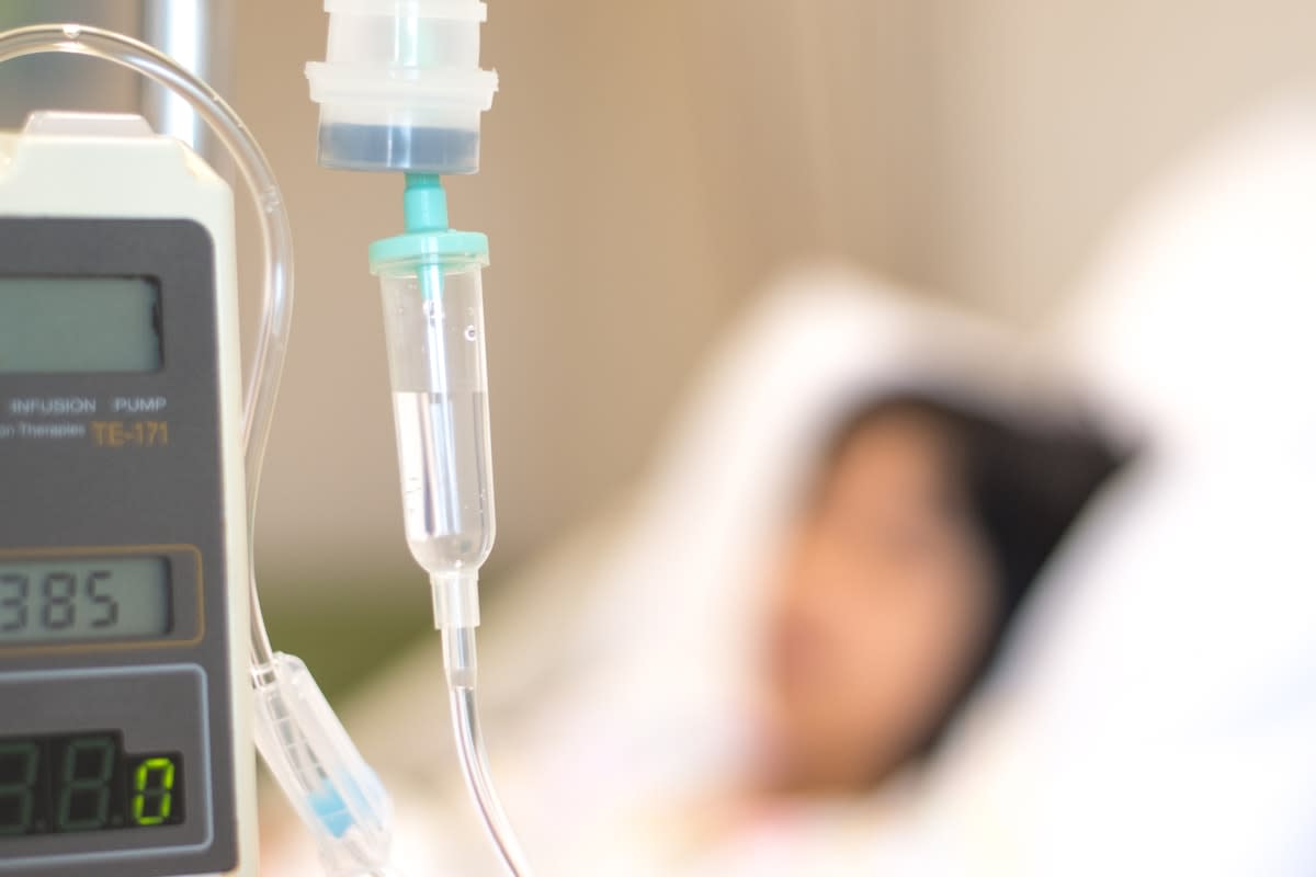 Close-up of an intravenous drip machine, with a blurred figure in a hospital bed in the background