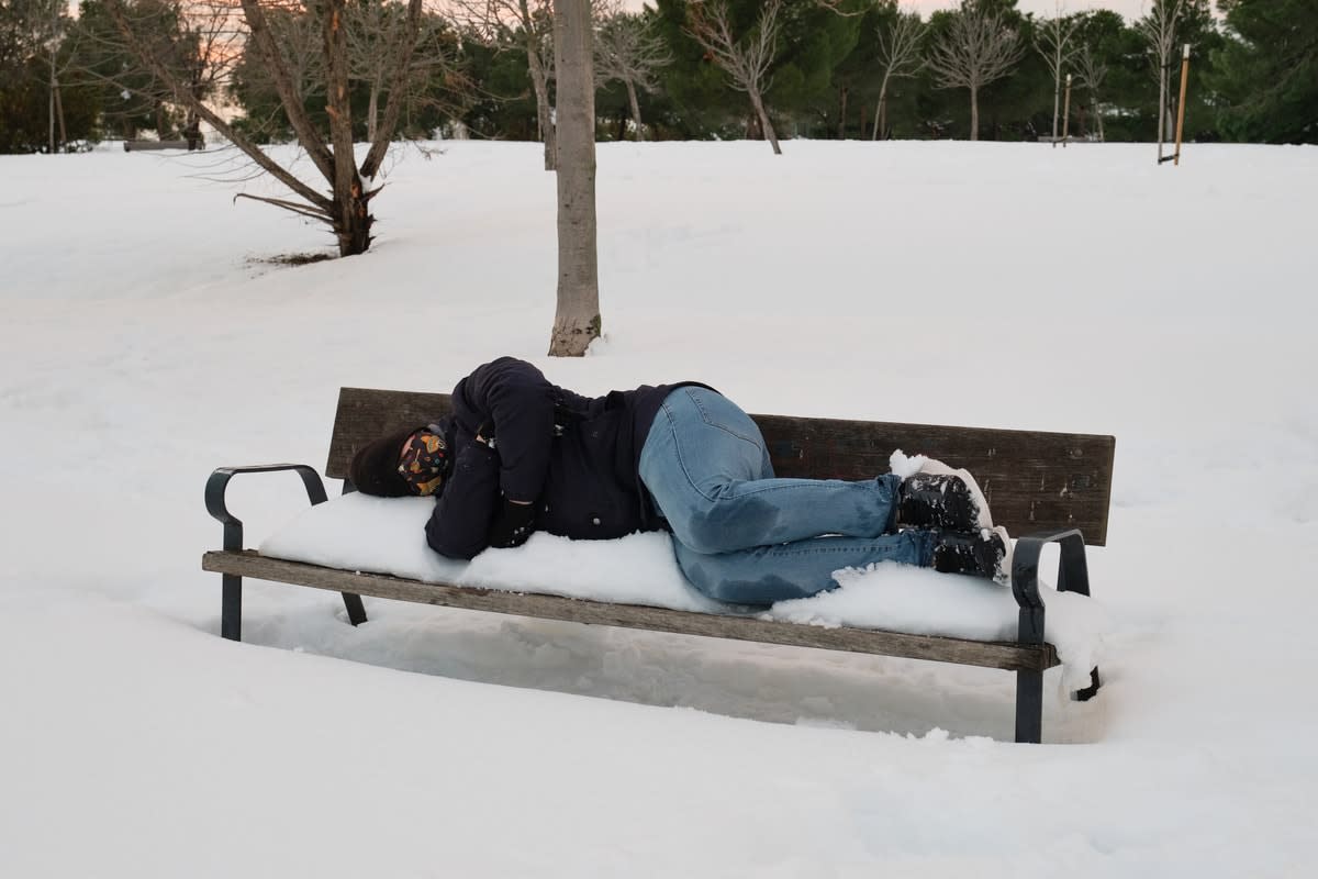 A homeless man asleep on a snow-covered park bench, surrounded by snow