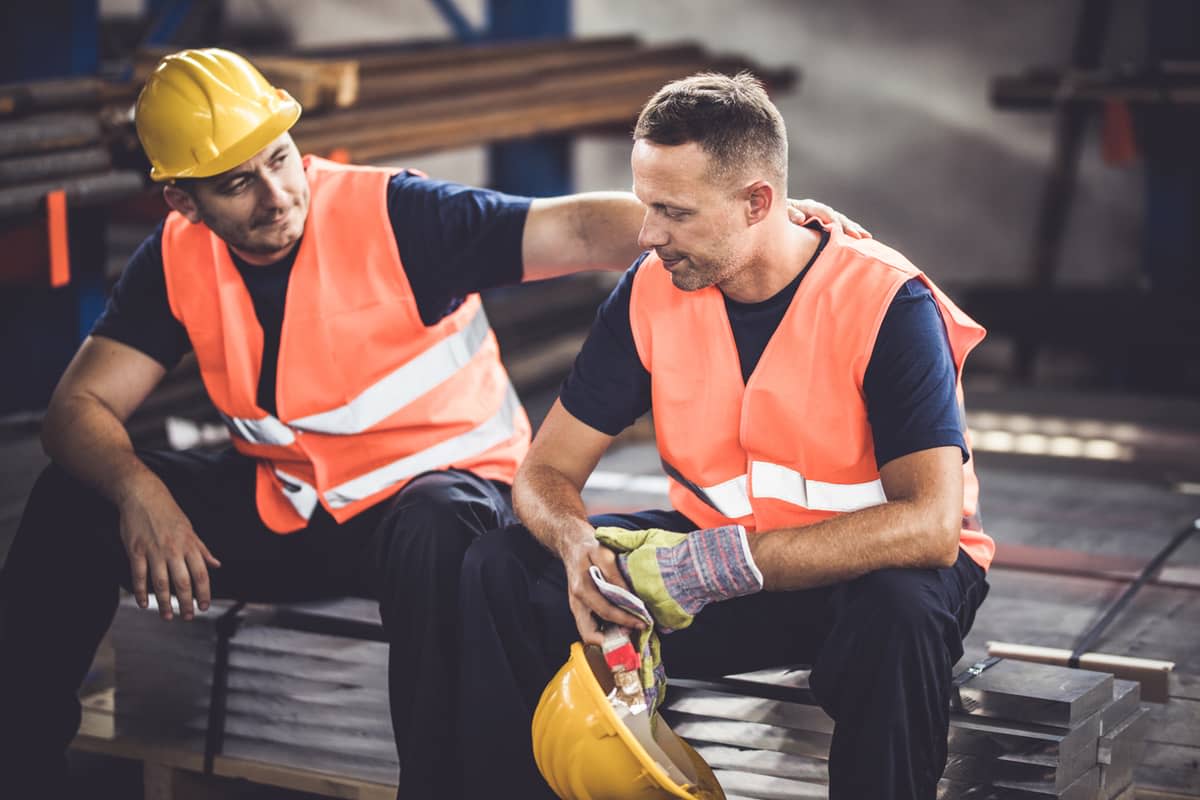 A construction worker wearing a hard hat sitting with a colleague and putting a comforting arm on his shoulders