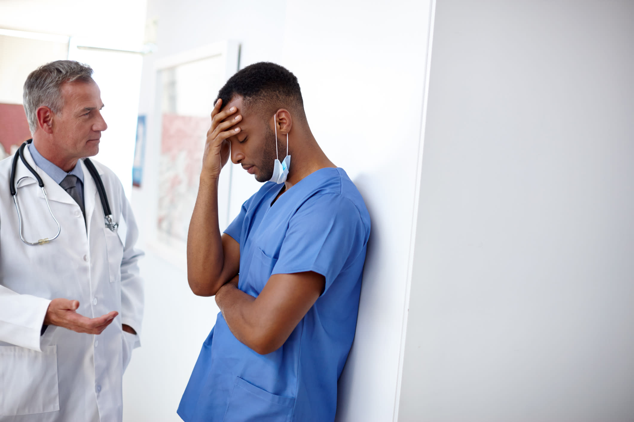 An older doctor in a white coat talking to a younger doctor leaning against a wall with his head in his hand