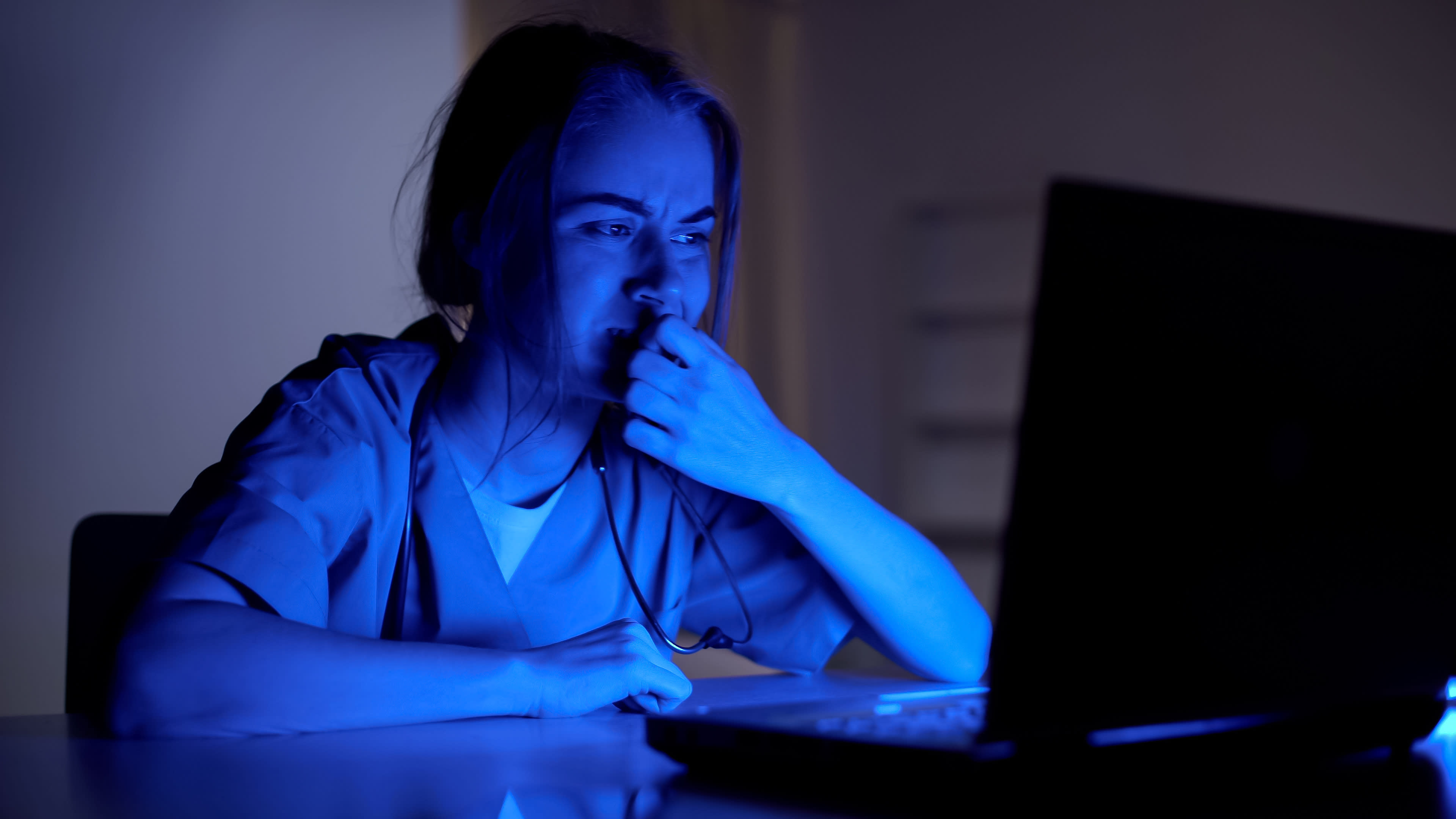 A stressed young female doctor stares at her laptop in a darkened room