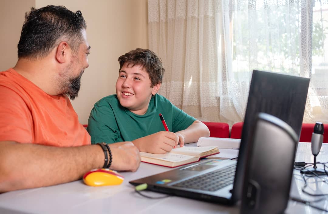 A boy and his father sit at a desk with a computer.