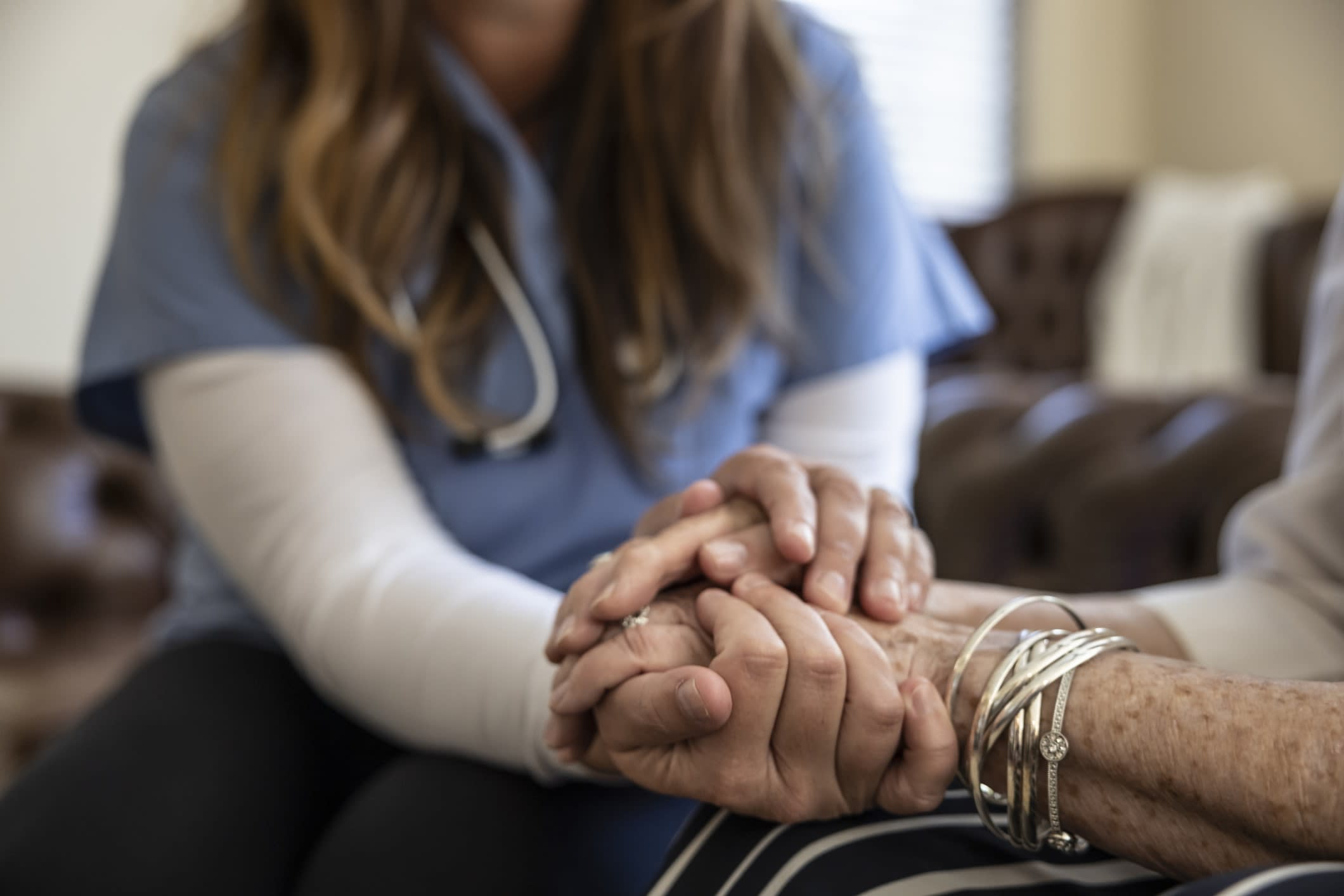 A seated female medical professional with a stethoscope around her neck, holding an elderly person's hand