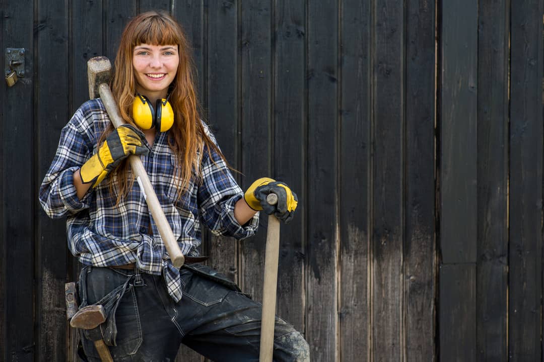 red haired girl in construction worker clothes standing with a shovel and a hammer