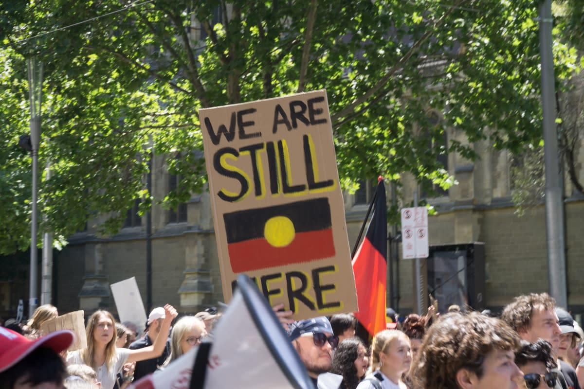 Protest marchers, one with a sign with the Aboriginal flag, reading ‘We are still here‘ 