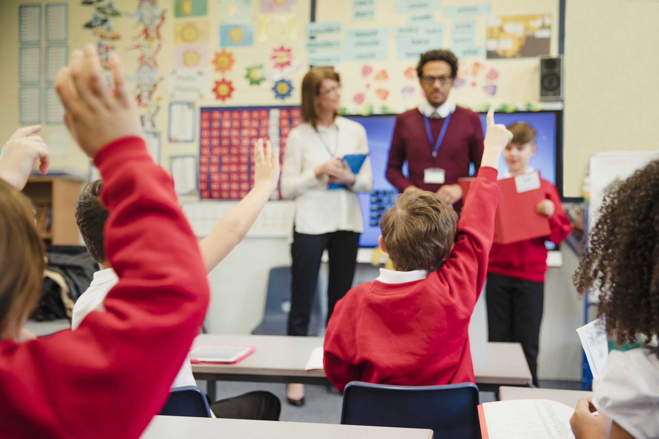 Students in classroom with their hands up, facing a student and teachers