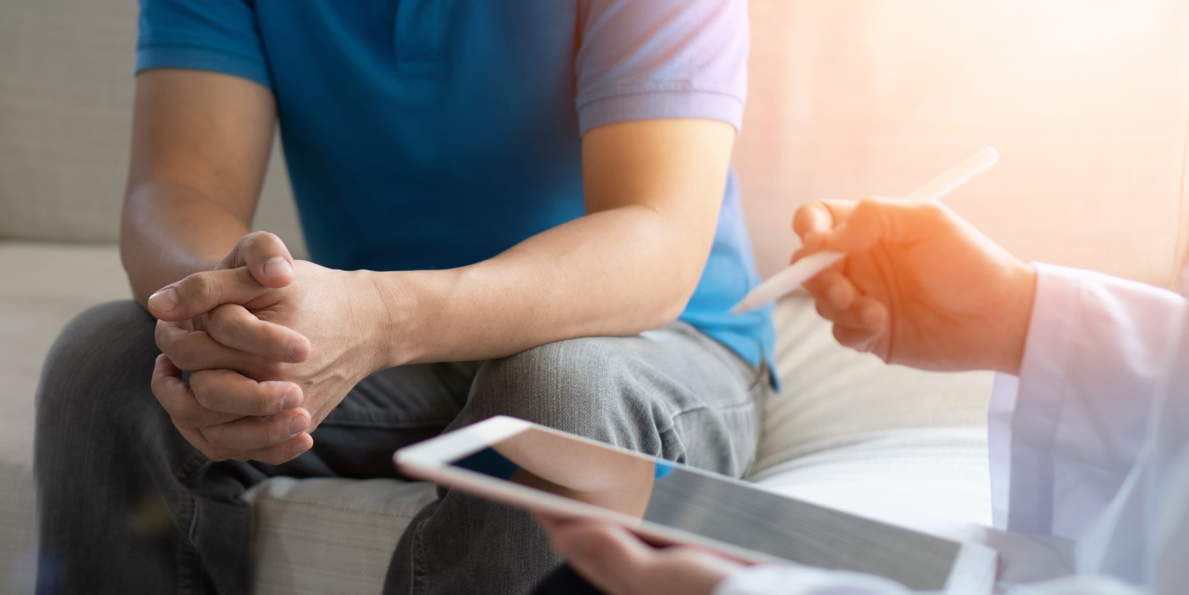 A seated male with clasped hands with a doctor, also seated, holding an iPad and stylus