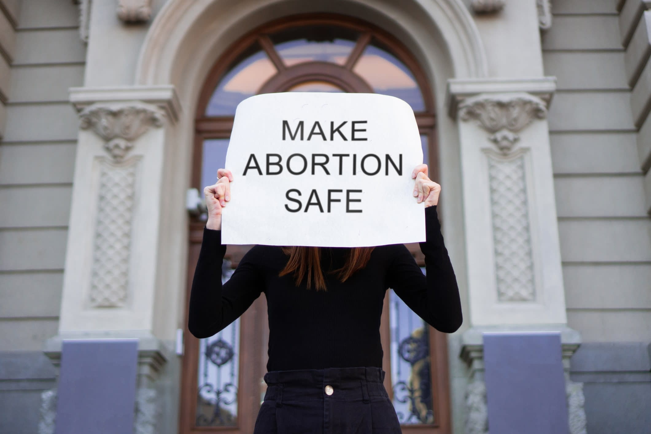 A young woman outside a courthouse holding a sign reading 'Make abortion safe'