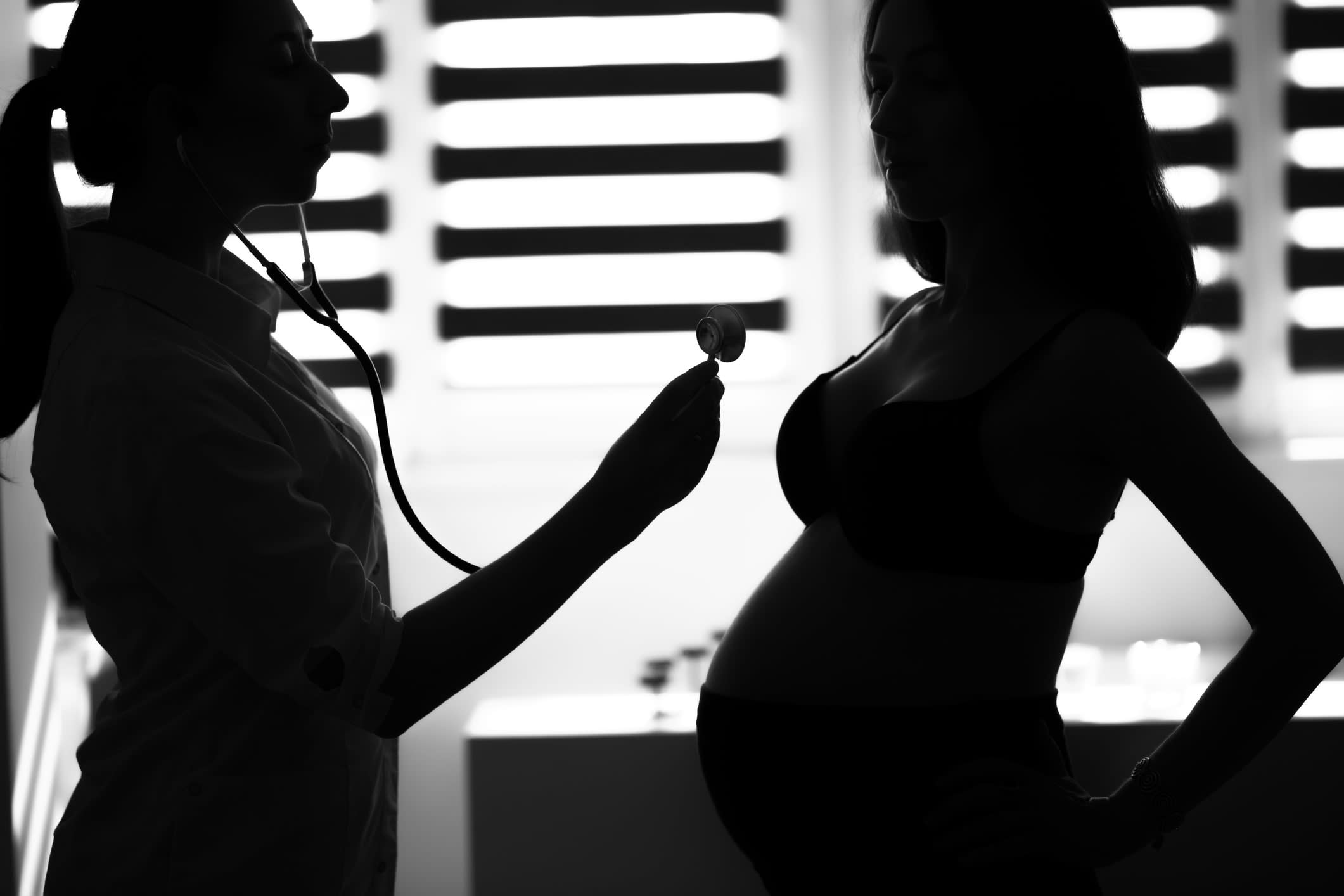 Black-and-white image of a silhouette female medicine doctor holding stethoscope to standing pregnant woman