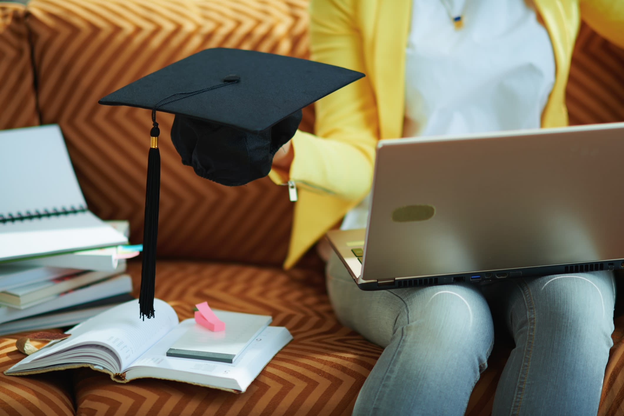 Woman sitting on a couch with a laptop, holding a graduation cap in on one, and books beside her 