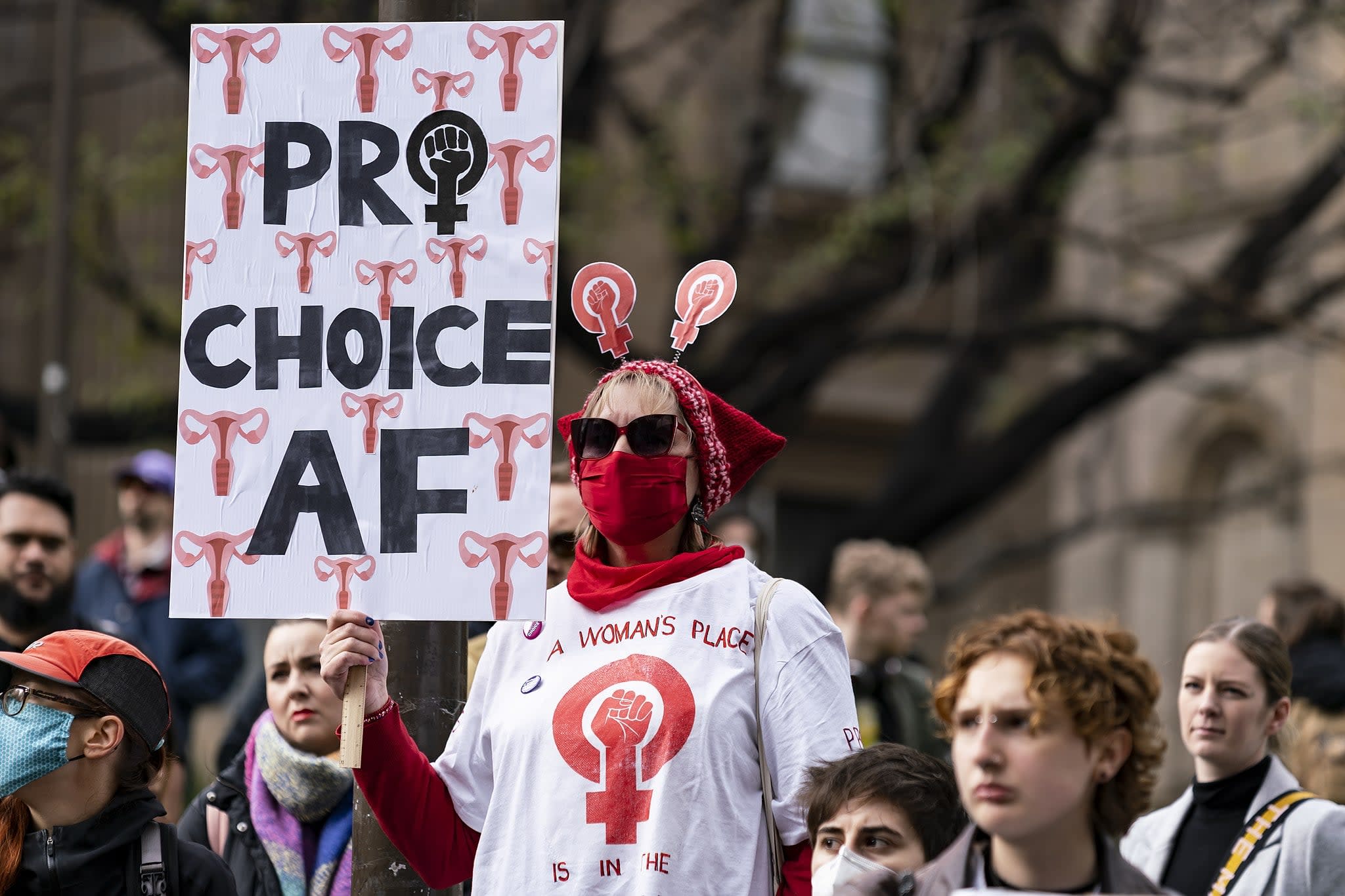 A woman wearing a red bandana on her face holding a sign at a protest reading 