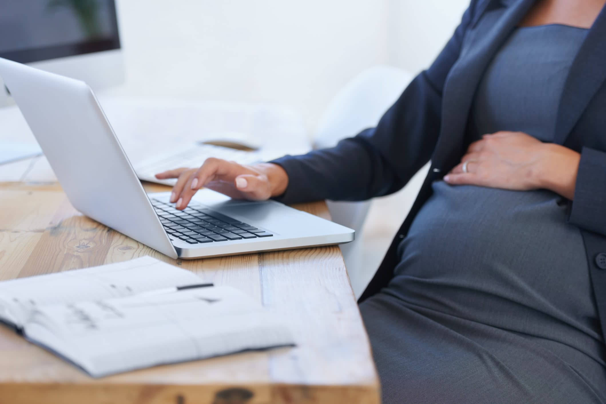 A pregnant woman in business attire sitting with one jhand on her stomach, the other typing on a laptop