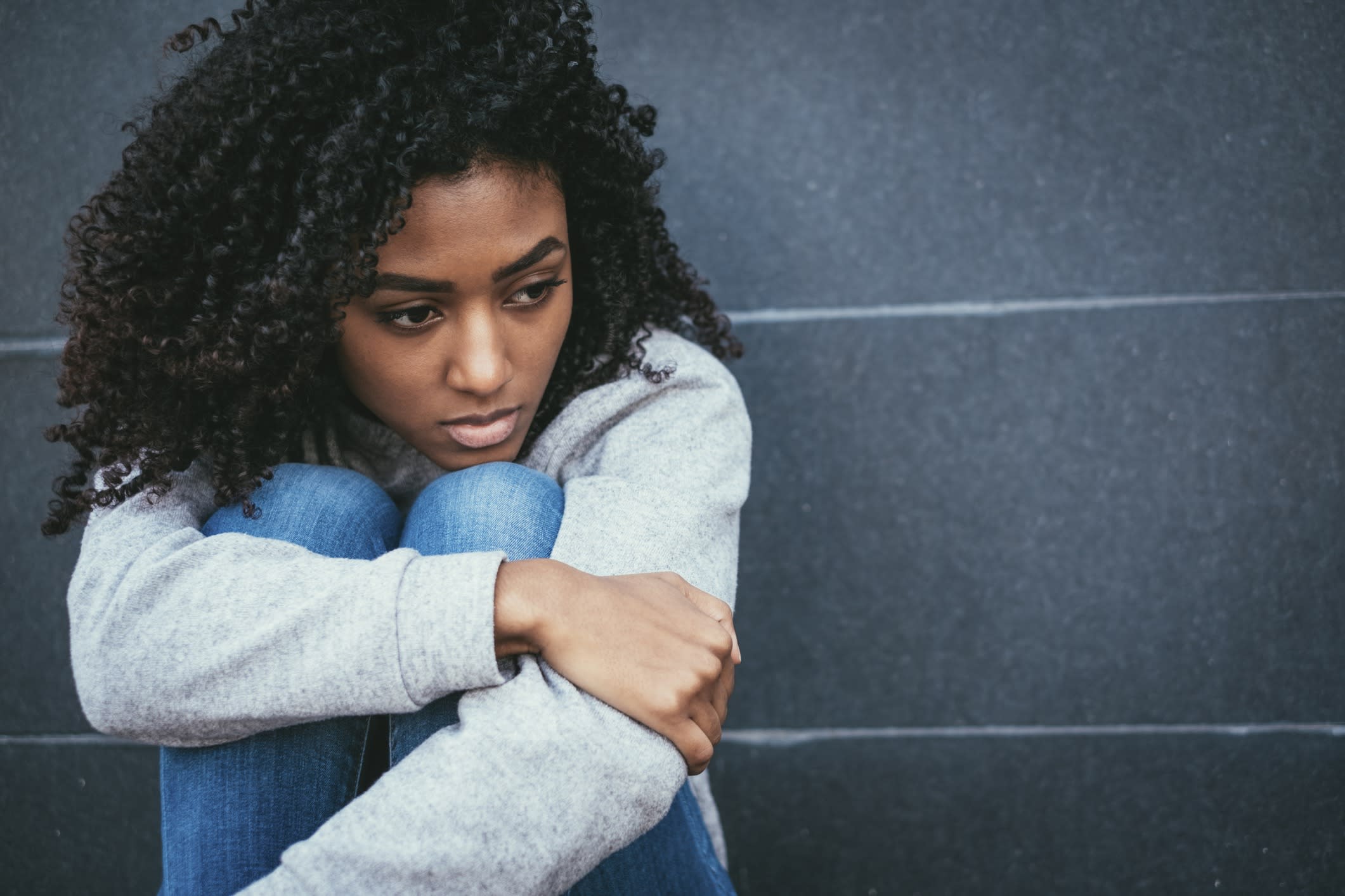 A young black woman sits alone with her knees tucked up under her chin looking pensive.