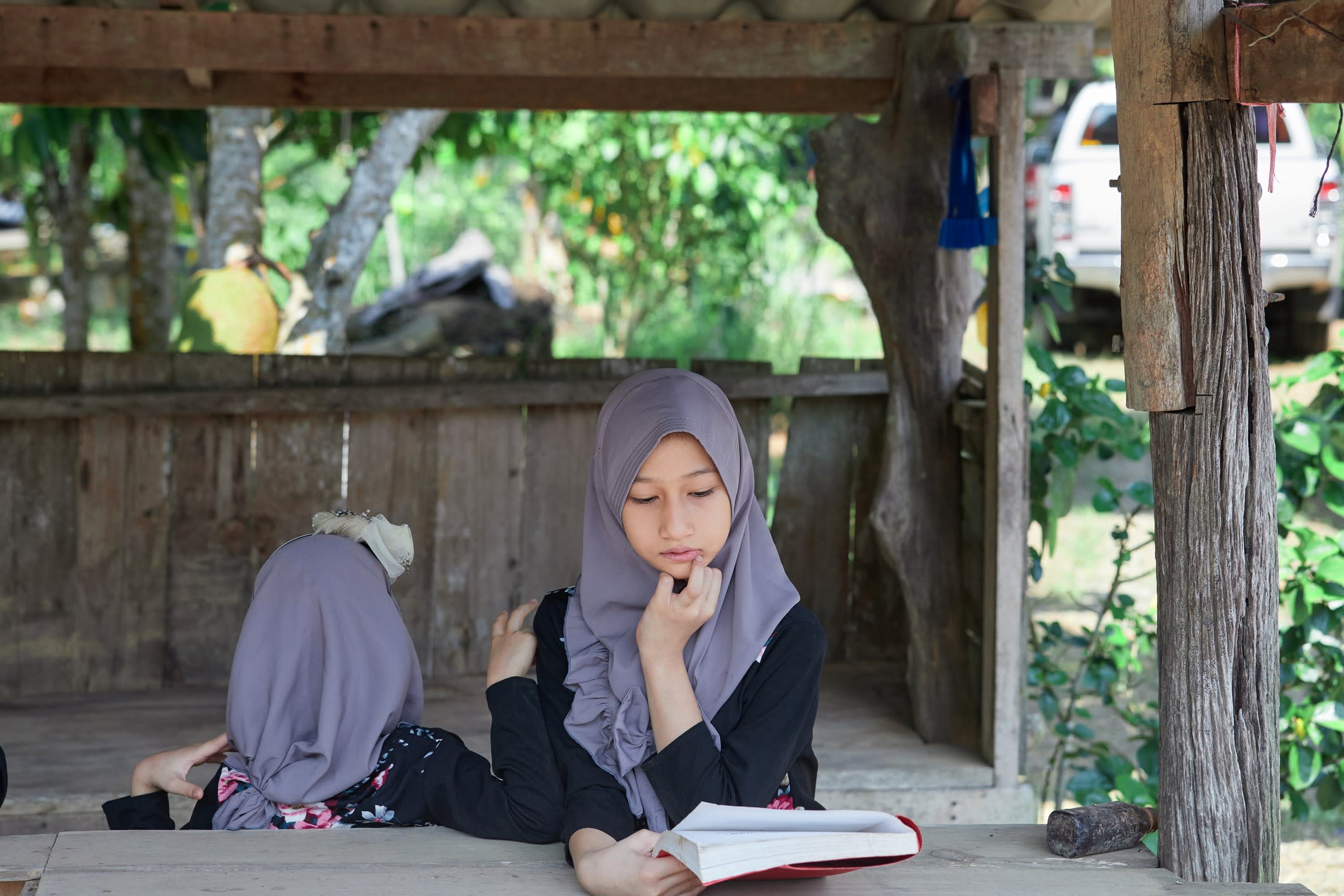 Asian Muslim girls wearing hijab and reading book at rustic pavilion