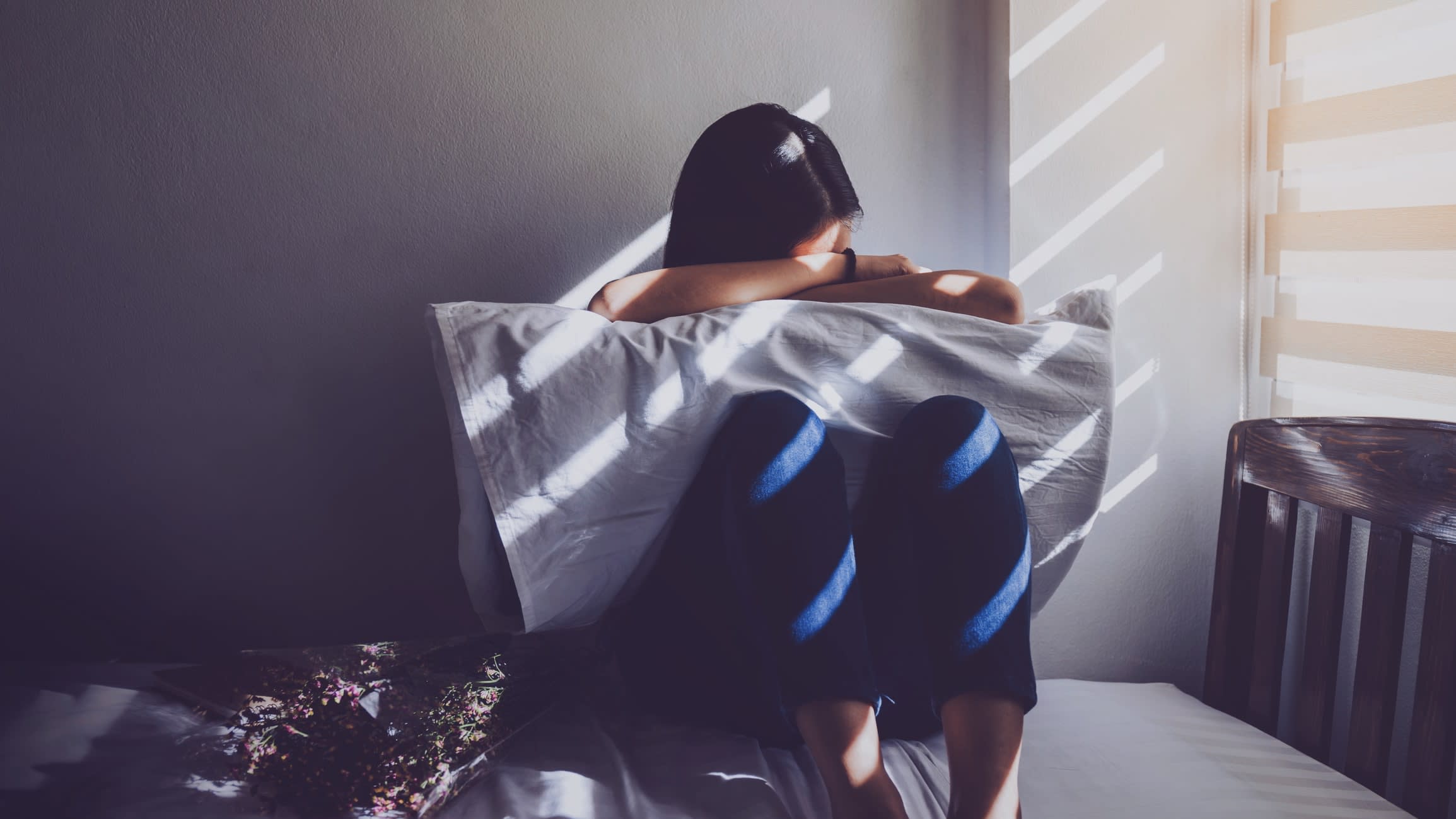 Women hugging a pillow and her knees on a bed in a dimly-lit bedroom, looking sad as sun shines in the window