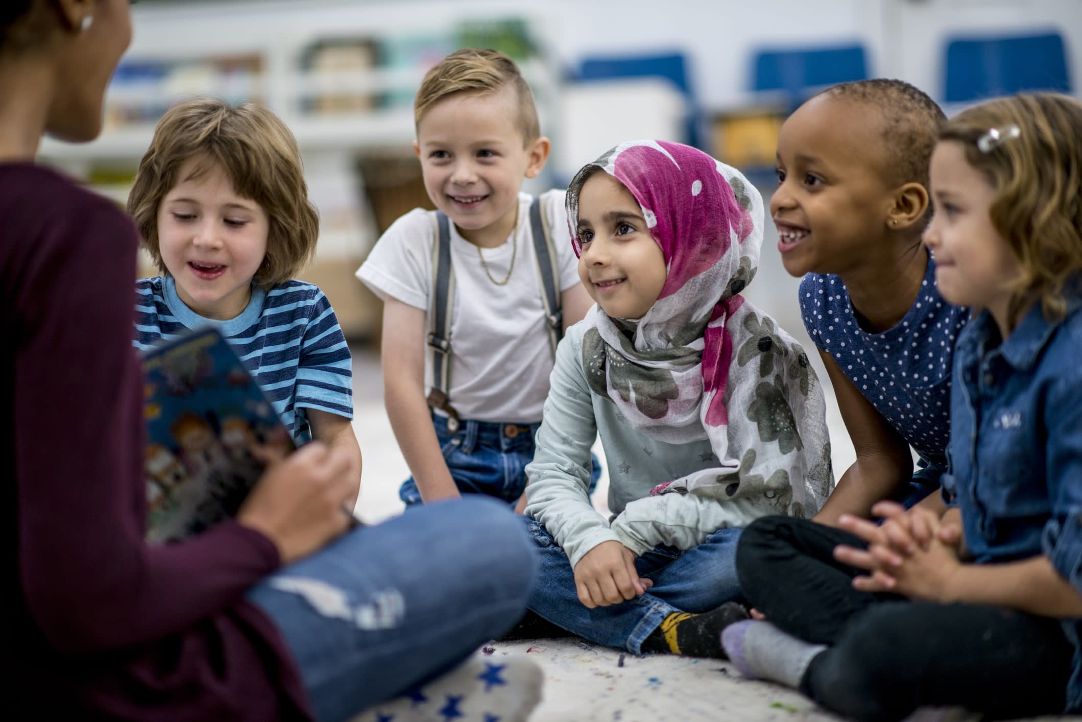 Smiling young children sitting on a classroom floor as the teacher reads a storybook.