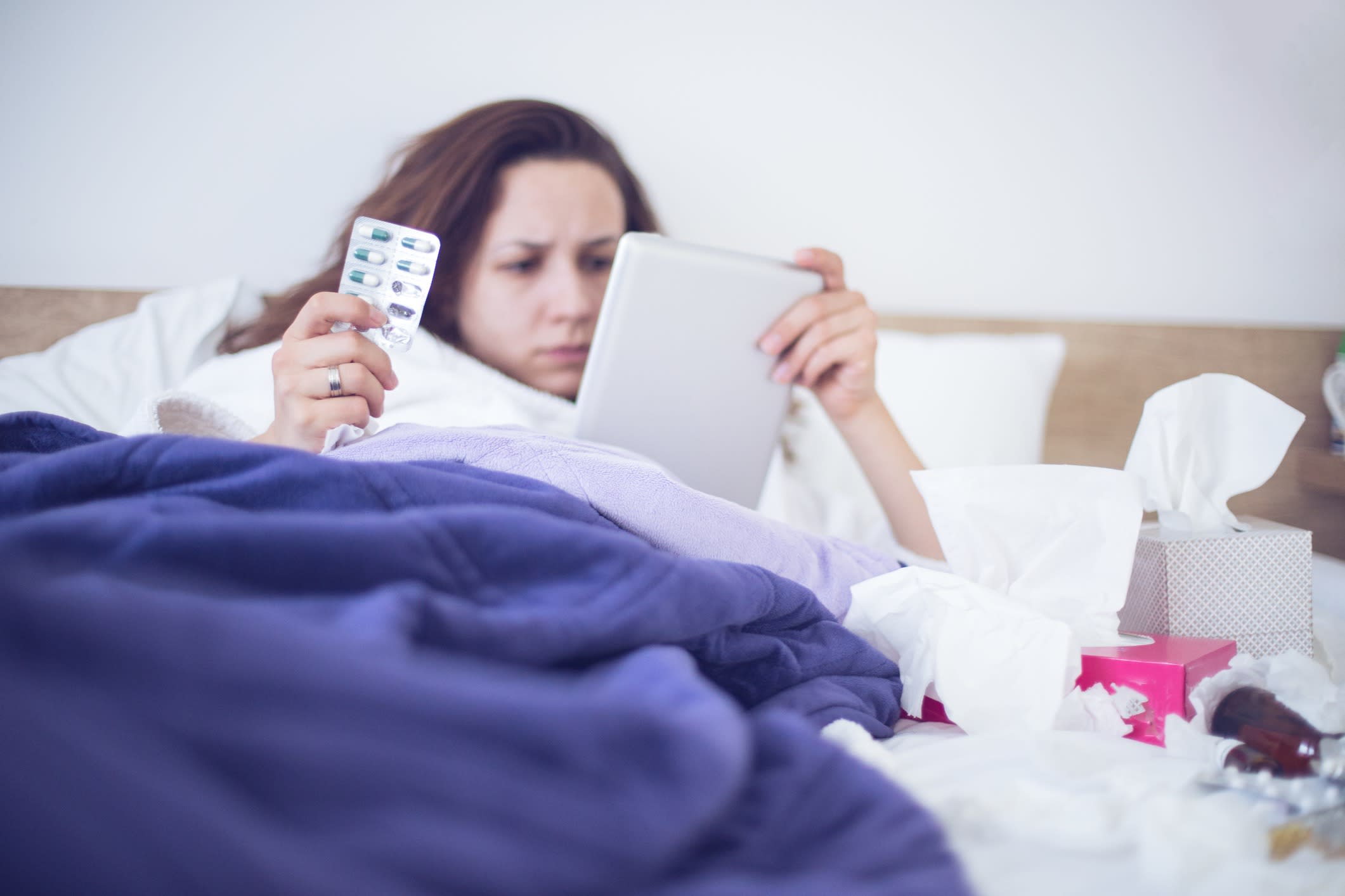 Ill woman lies in a bed searching on her computer for medical information.