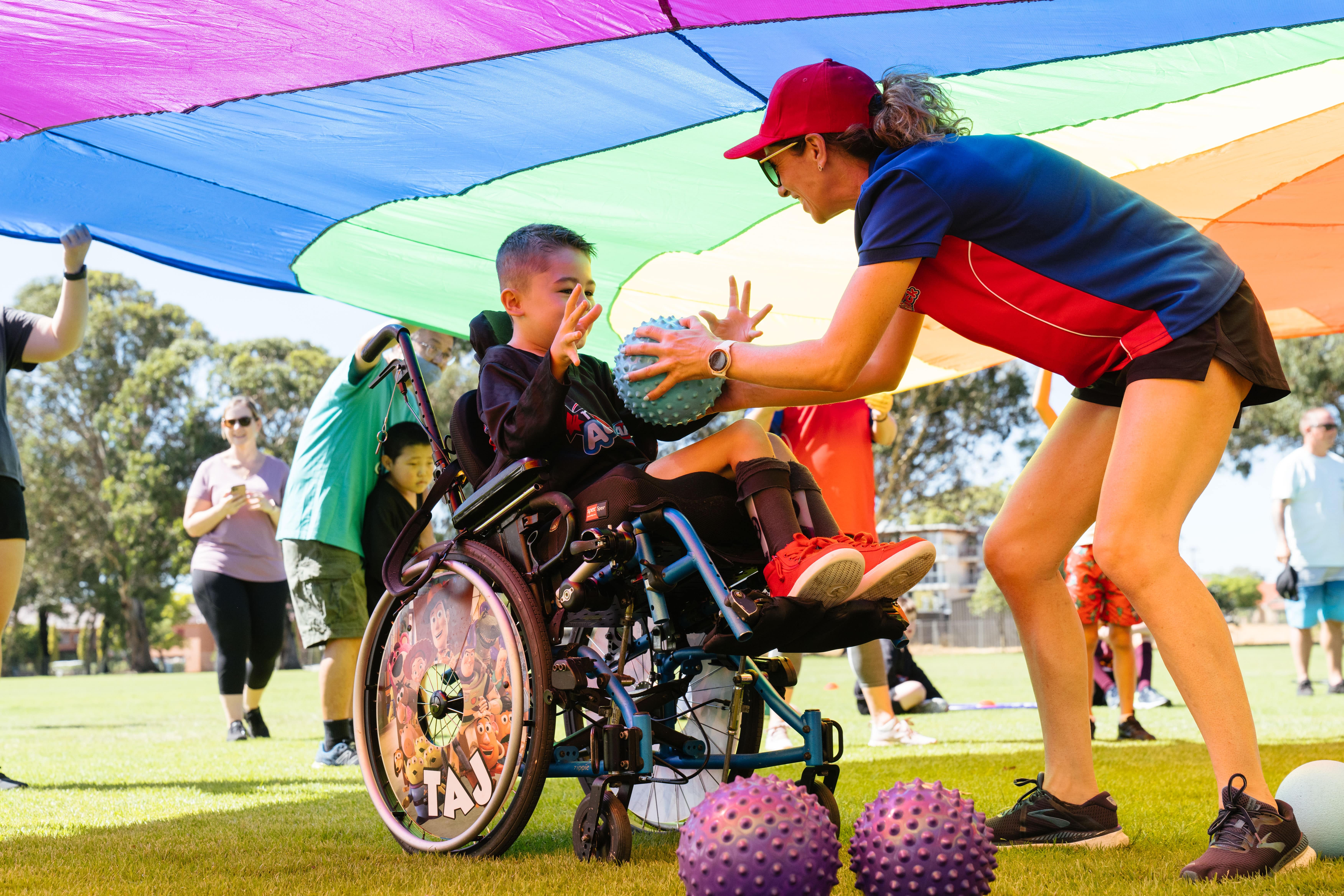 An instructor handing a ball to a child in a wheelchair under a colourful banner