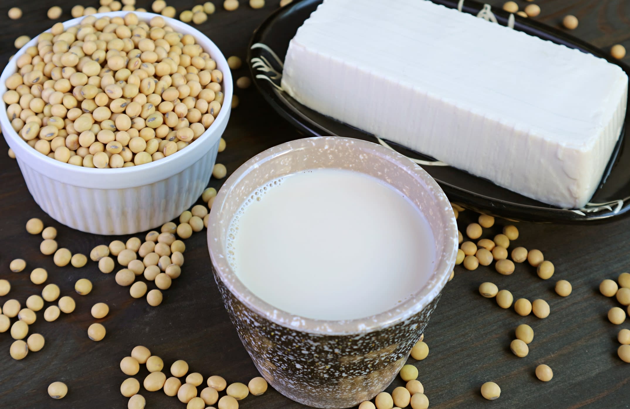 Cup of soy milk, soft tofu and dried soybeans in a container, with soybeans scattered around