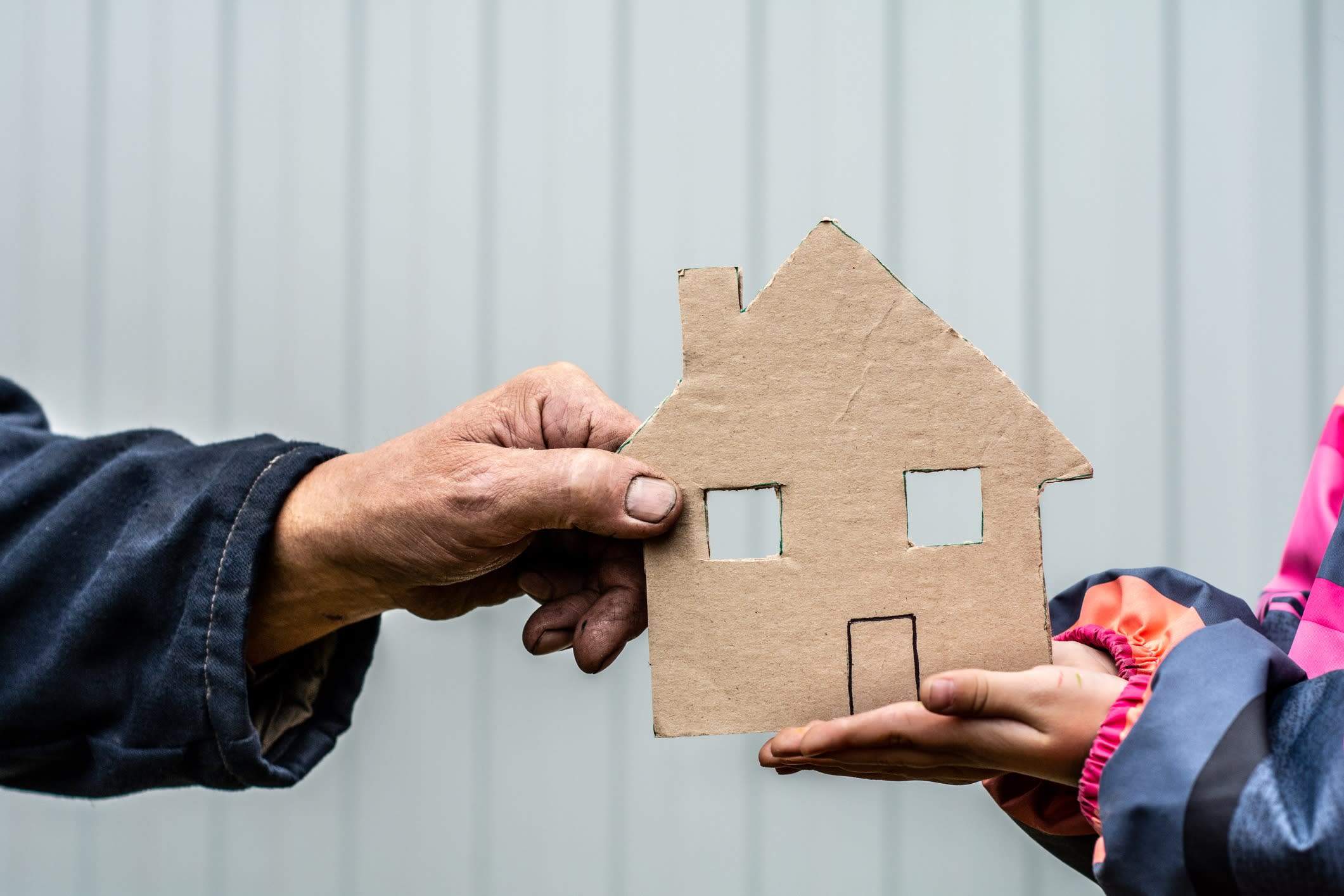 A man's hand holds a paper house cut out of cardboard. Nearby, children's hands hold the house 