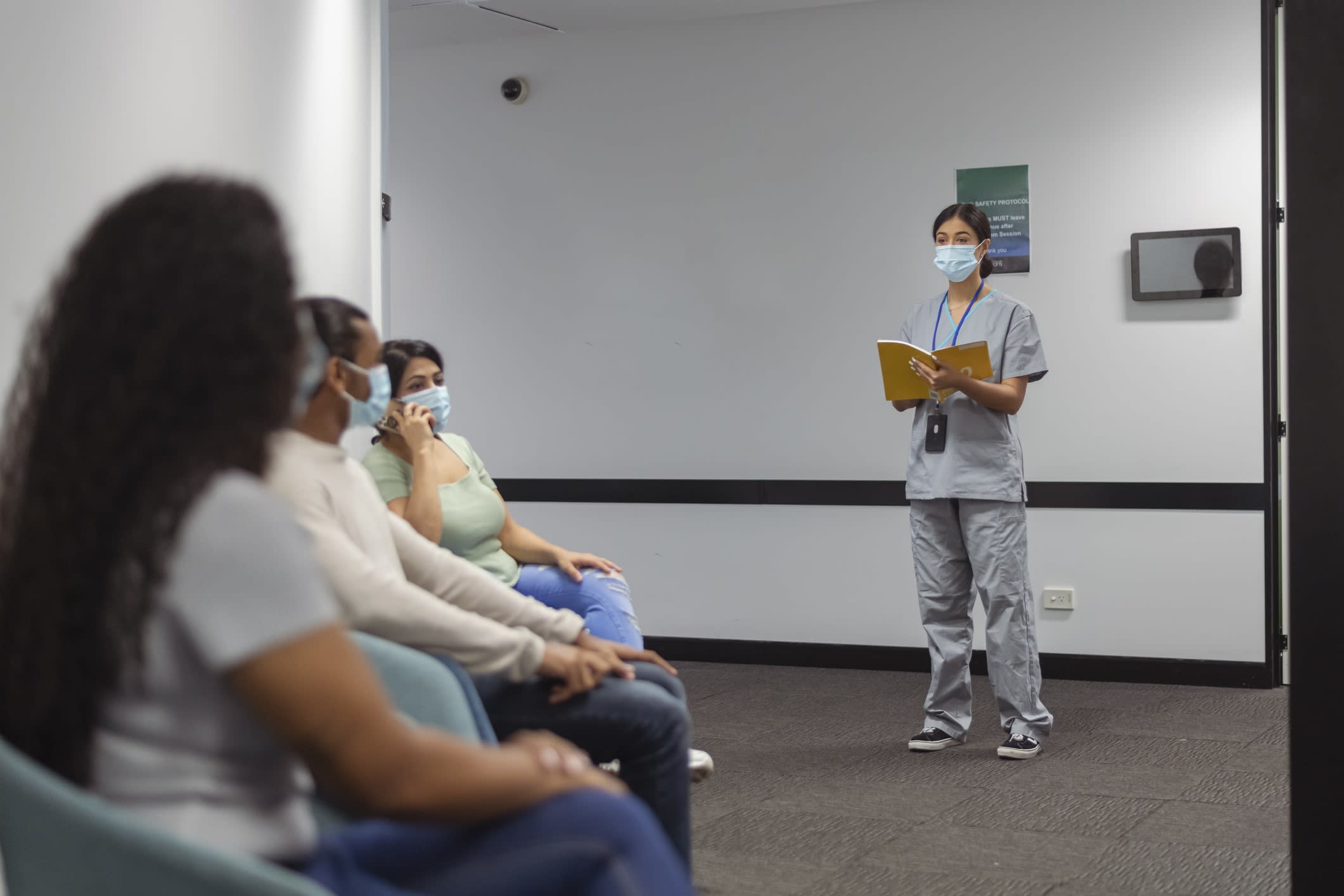 A masked nurse talking to seated patients in a hospital hallway