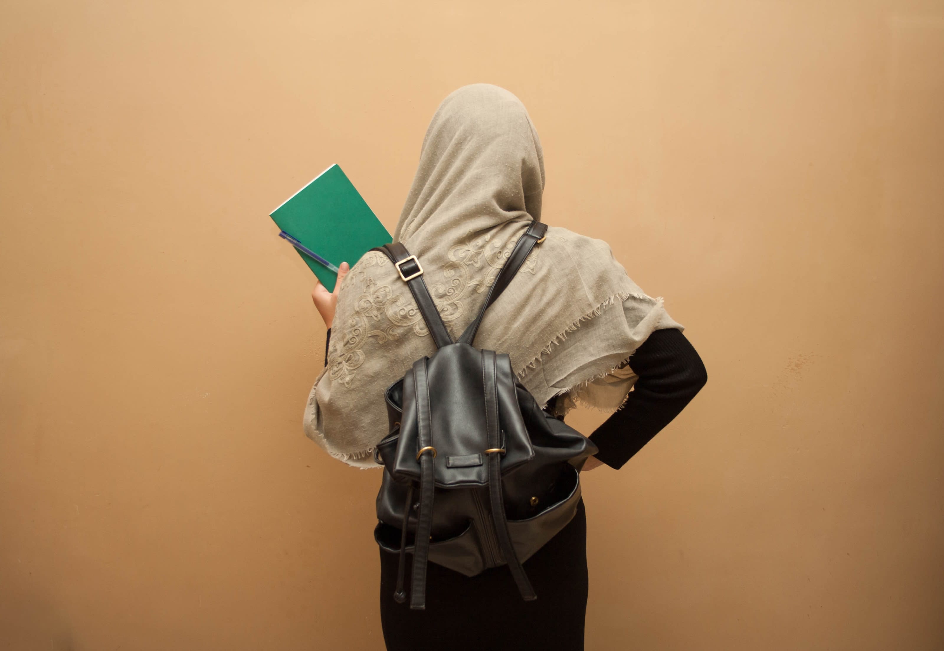 A Muslim schoolgirl wearing a backpack and holding a book, facing away from the camera