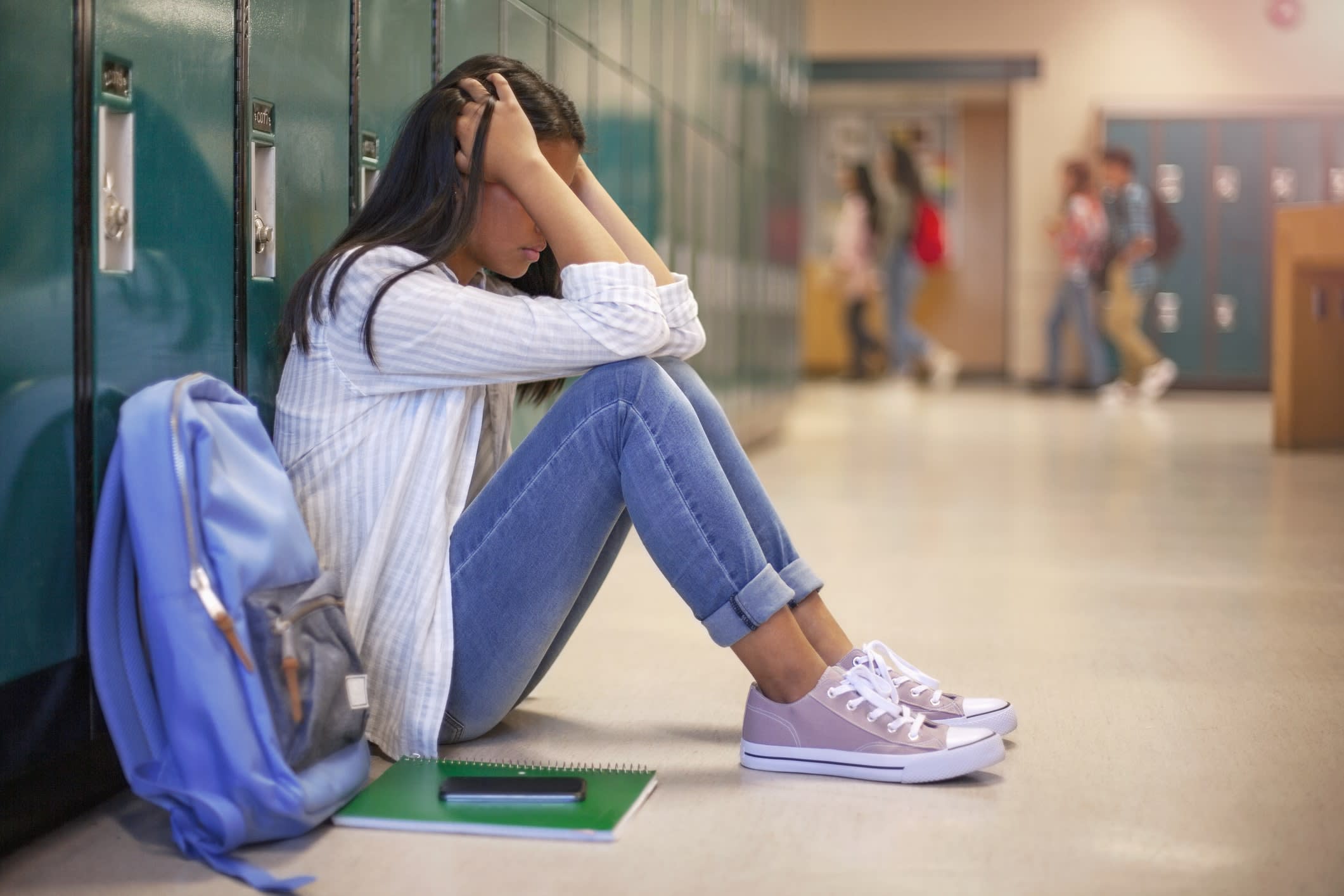 A depressed girl sits in a school corridor with her back to lockers and her head in her hands