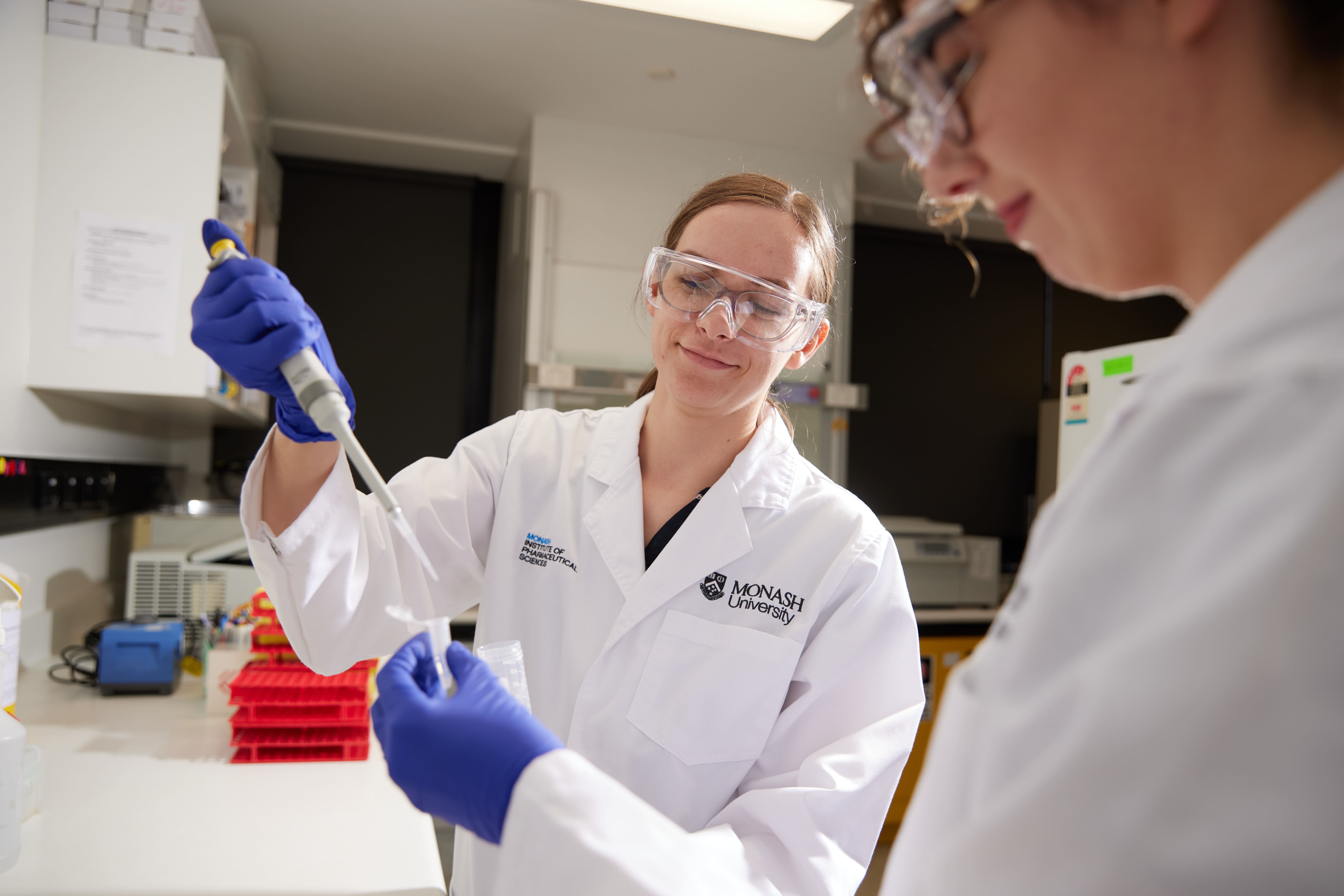 Dominika Fuhs, in protective goggles and lab coat, adds liquid to a test tube with the help of a colleague