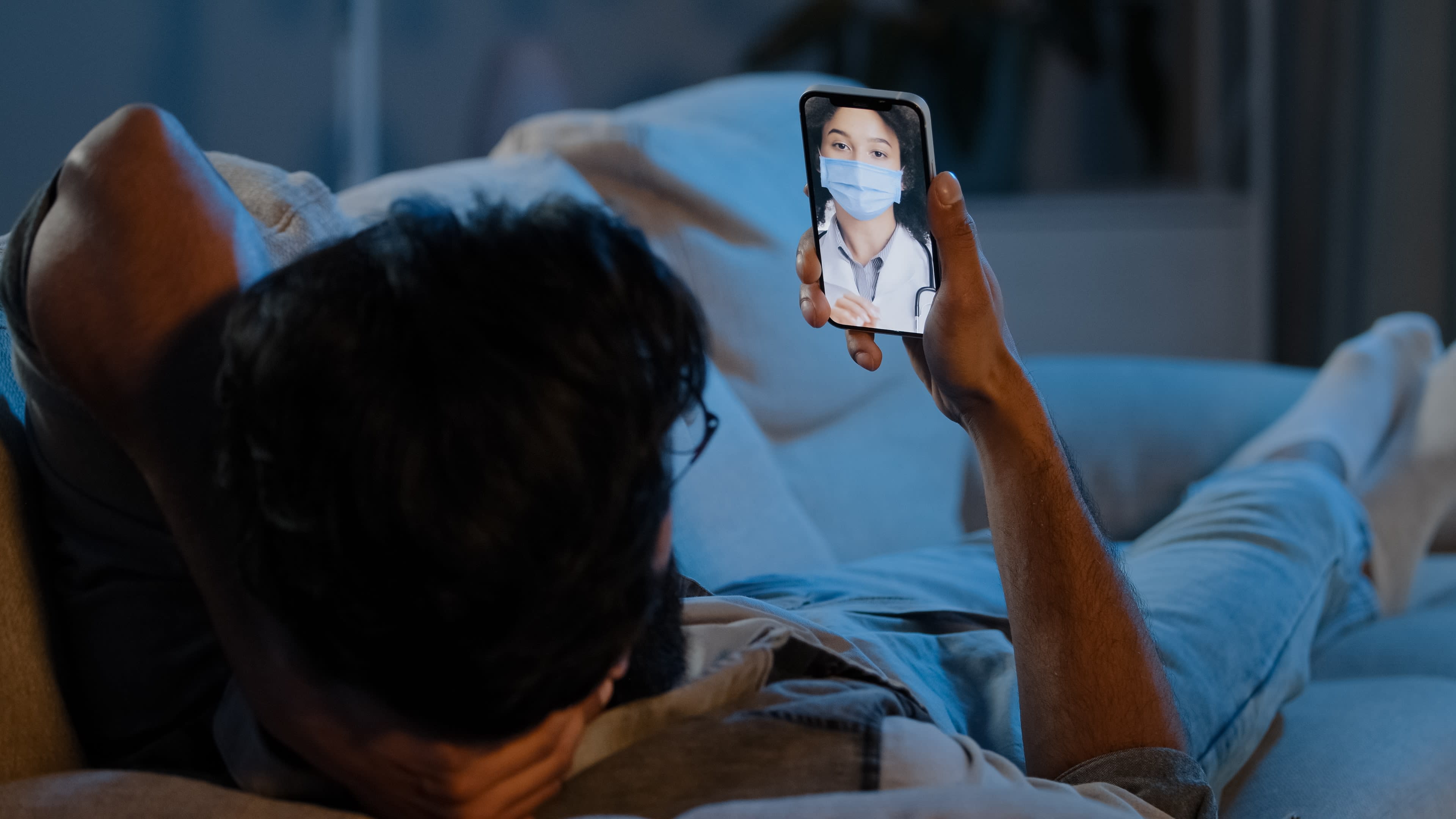 Man lies on couch with his phone talking to a masked doctor in a telehealth consultation video call