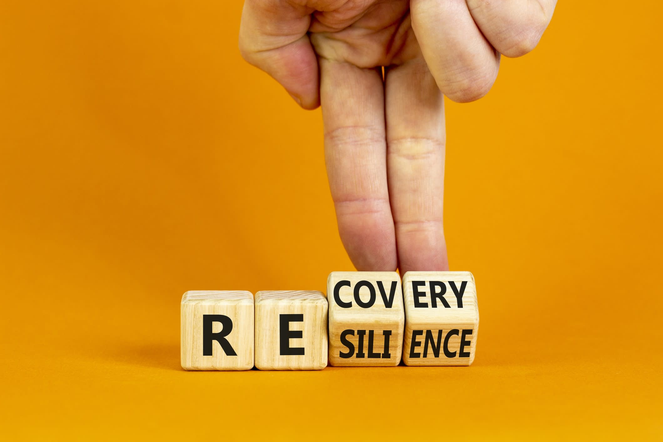 Fingers turning cubes and changing the word 'recovery' to 'resilience' on an orange background.