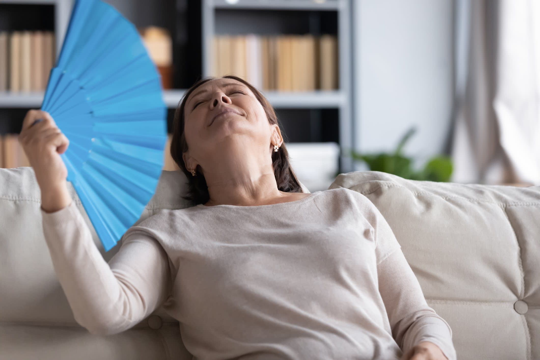 A middle-aged woman on a couch leaning back and fanning herself with a blue fan