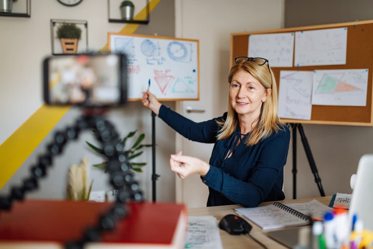 Image of a female teacher sitting in an office facing a camera phone, teaching remotely