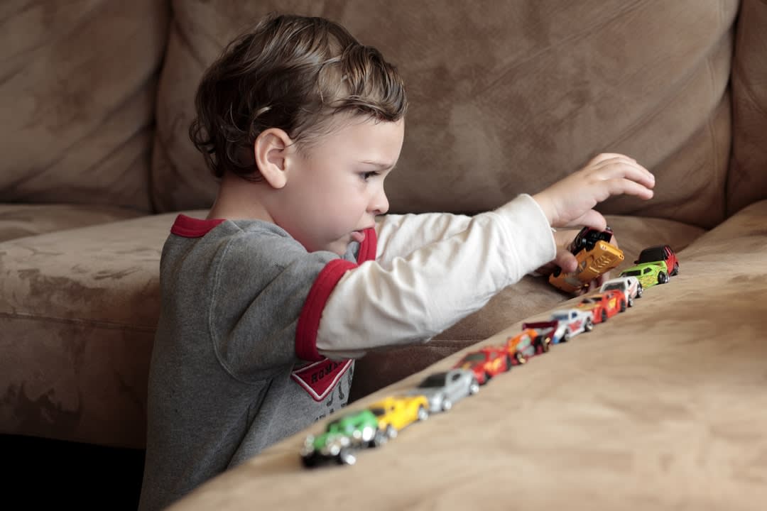 A young child plays with a row of cars on a brown sofa.