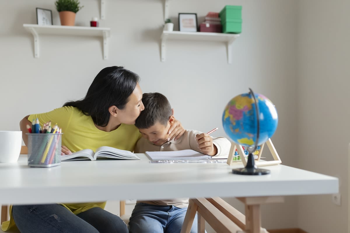 Picture of a mother kissing her young son, who's holding a pencil, on the head while sitting at a desk during home schooling