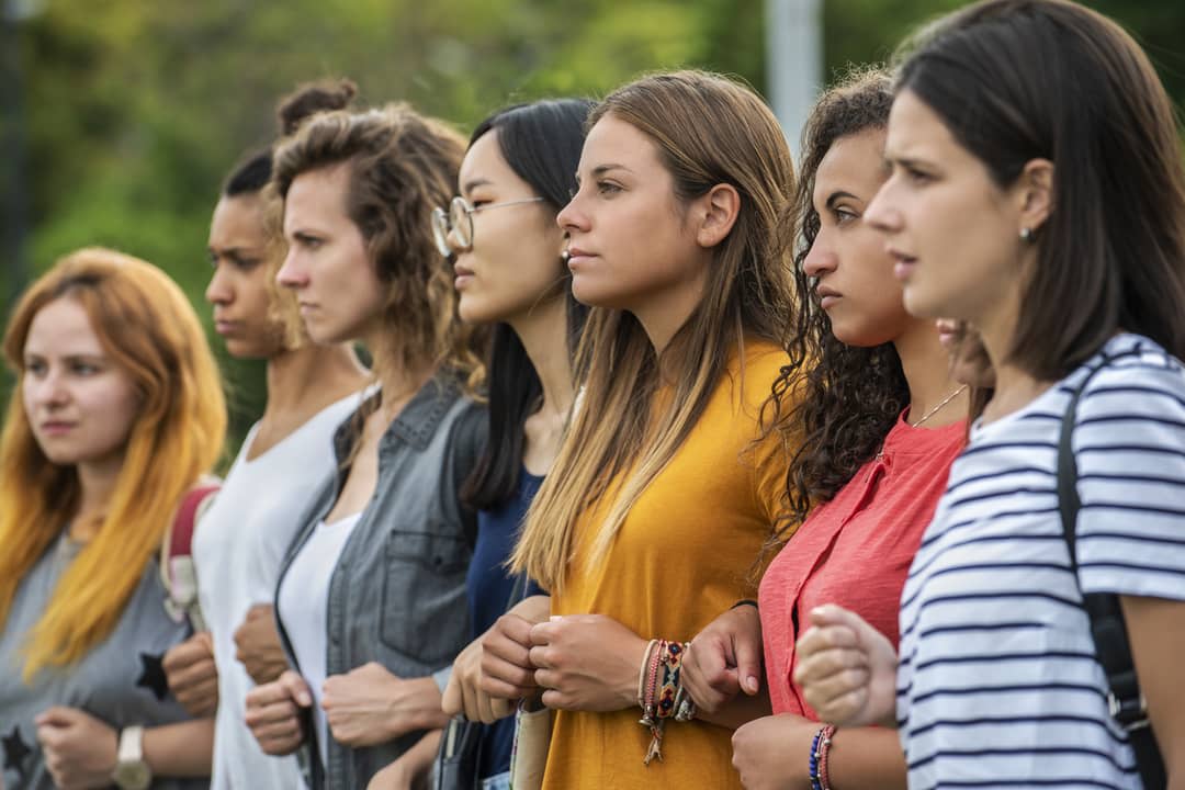 A row of determined women stand in a line locking arms.