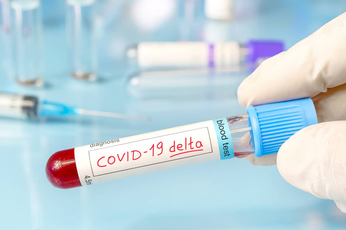 A latex-gloved hand holding a blood-test vial with a label reading ‘COVID-19 Delta‘