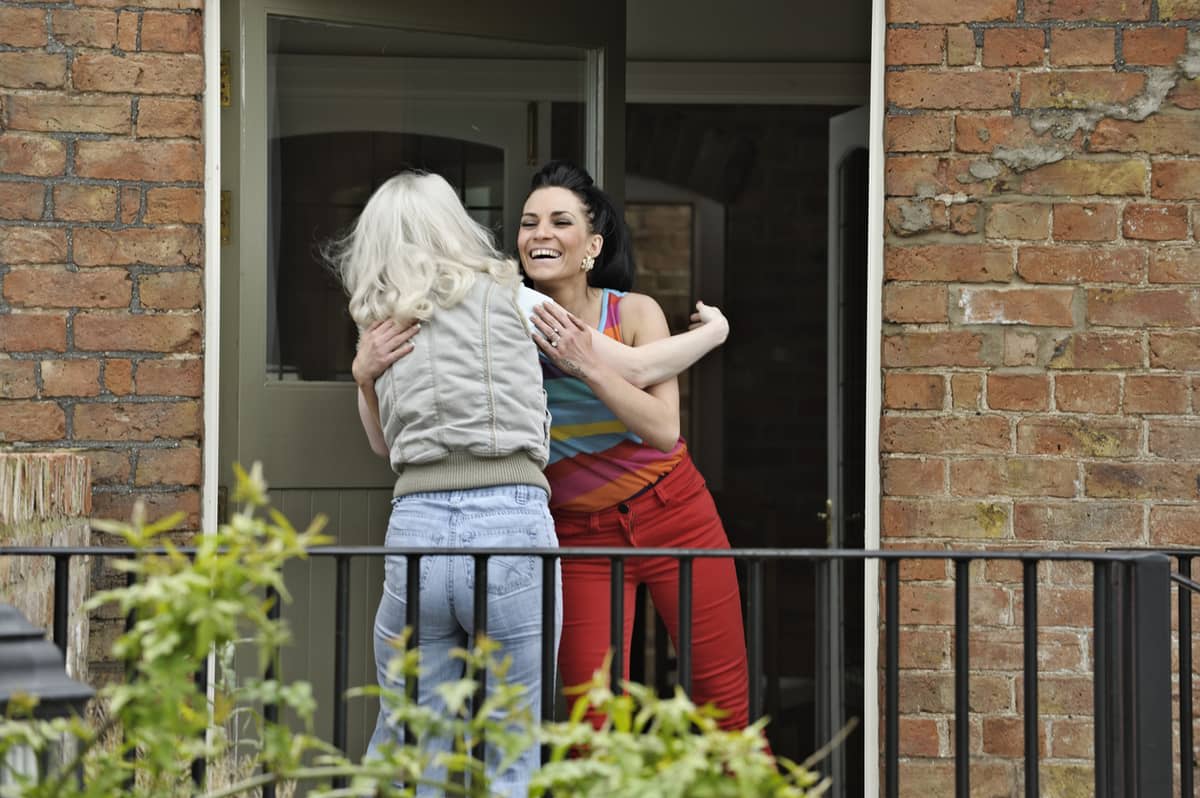 A smiling women greets another woman with a hug in a doorway