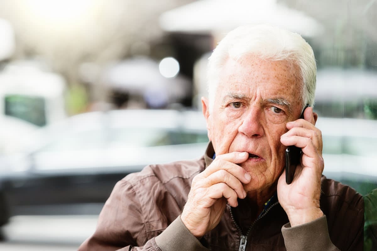 A worried-looking older man with his hand to his mouth, holding a mobile phone