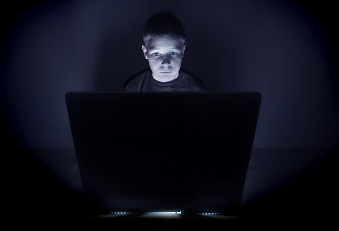 A frightened child sits in front of a computer screen.