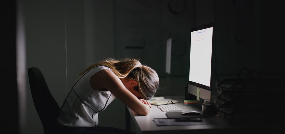 A woman sits at her desk in front of her computer with her head in her hands.