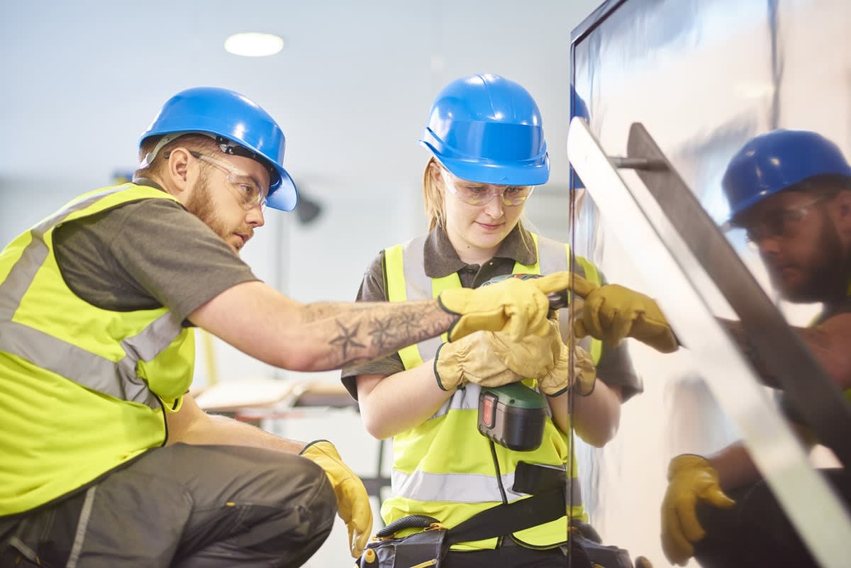 A woman in hard hat and high-vis clothing being instructed by a male dressed the same on using a drill on a construction site. 