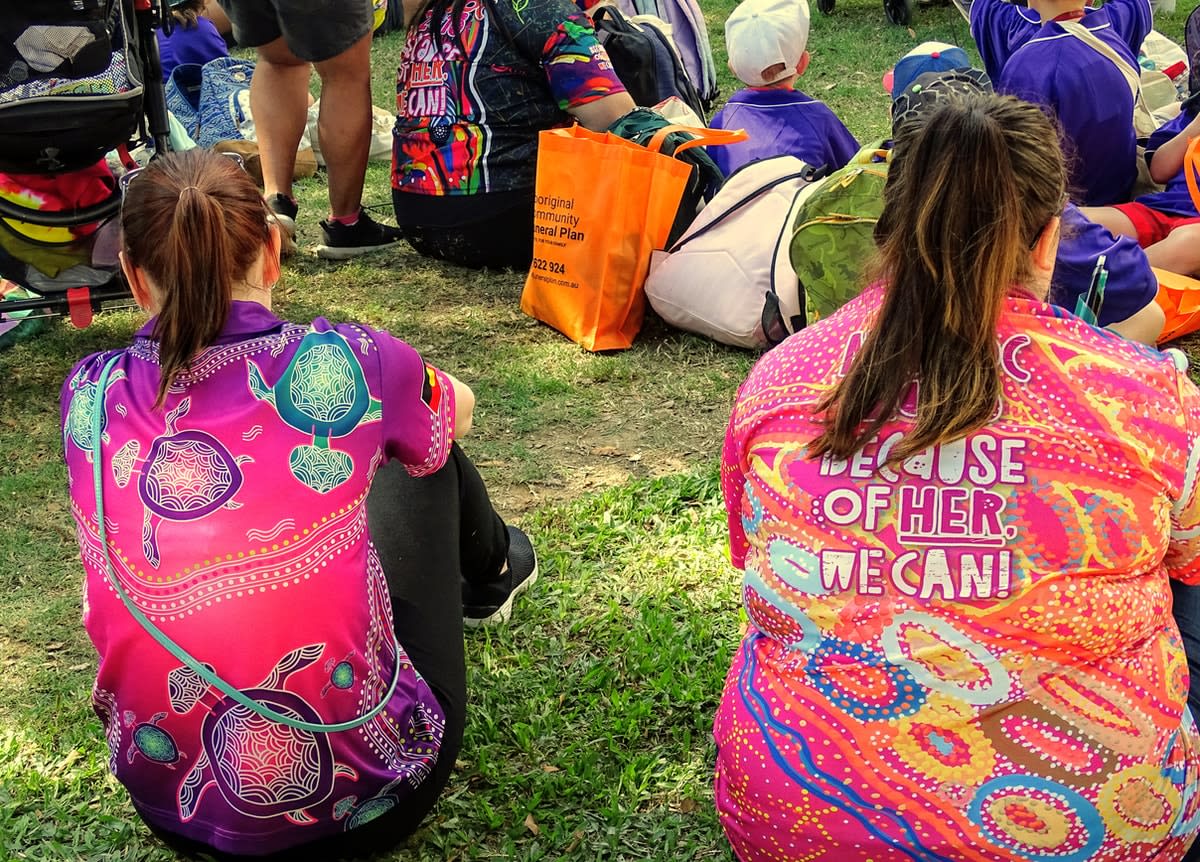 Two women wearing Indigenous design shirts, photographed from the rear, sitting on the grass among a crowd