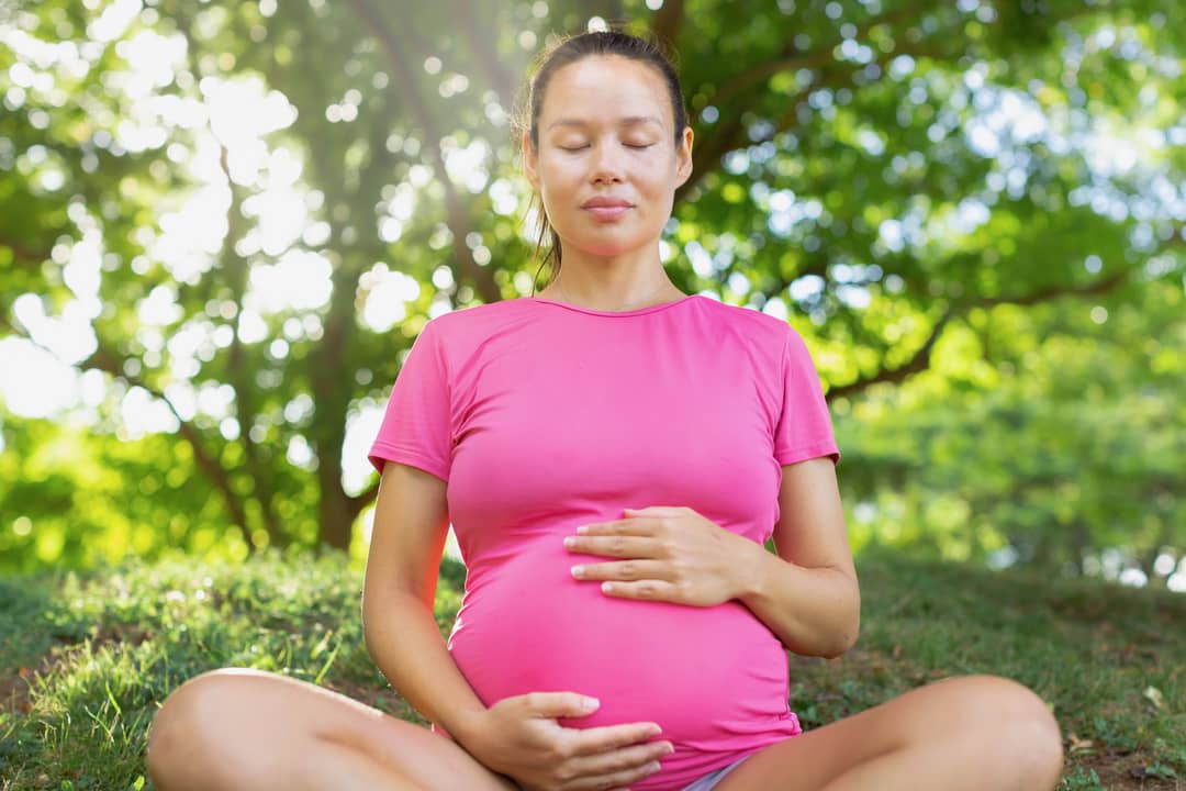 A pregnant woman sits in a park surrounded by greenery.